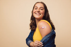Portrait of a female smiling after getting a vaccine. Woman holding down her shirt sleeve and showing her arm with bandage after receiving vaccination.